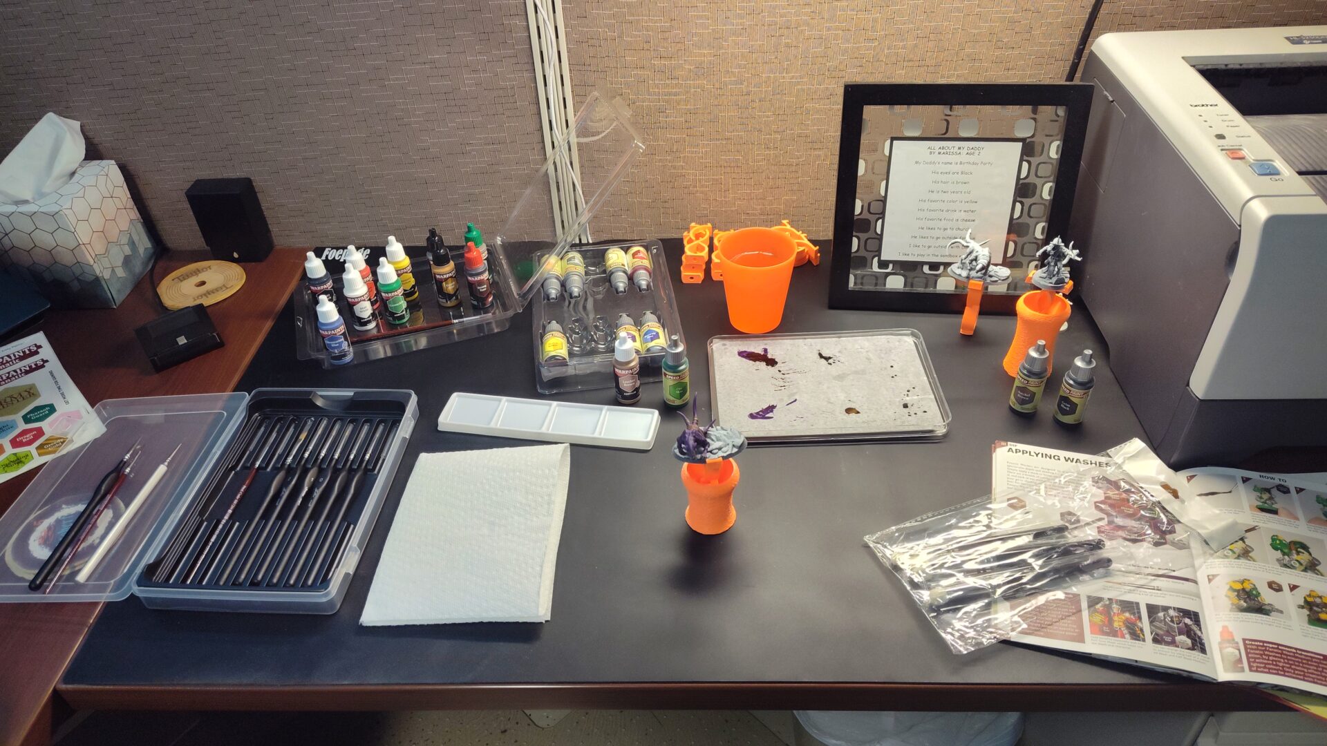 Painting station setup with black rubber mat, Army Painter paints, water for cleaning brushes, and some miniatures ready for painting.